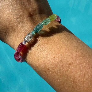 Könnte beinhalten: Ein mehrfarbiges Perlenarmband mit verschiedenen Farbtönen, darunter Rosa, Blau, Grün und Klar. Das Armband wird an einem Handgelenk getragen, mit einem blauen Hintergrund. Die Perlen scheinen geschnitzt oder strukturiert zu sein.