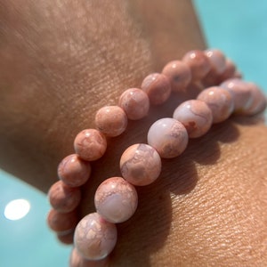 Puede incluir: Dos pulseras de cuentas de ágata rosa y blanca apiladas en la muñeca de una persona. Las cuentas tienen un patrón de mármol.