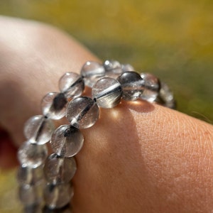 Puede incluir: Dos pulseras de cuentas con cuentas redondas y transparentes e inclusiones negras. Las pulseras se llevan en una muñeca, con un fondo natural borroso. Las cuentas tienen aproximadamente 1,3 cm de diámetro.