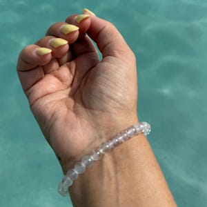 May include: A clear, beaded bracelet is worn on a wrist. The bracelet is made of small, round, translucent beads. The hand has yellow nail polish. The background is a light blue, possibly water.