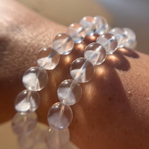May include: Two clear quartz crystal bead bracelets. The beads are round and translucent, strung together on a clear elastic cord. The bracelets are worn on a person's wrist, with sunlight casting shadows.