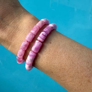 May include: Two pink beaded bracelets with a marbled effect are worn on a person's arm. The bracelets are made of cylindrical beads in shades of pink and white. The background is a bright blue.