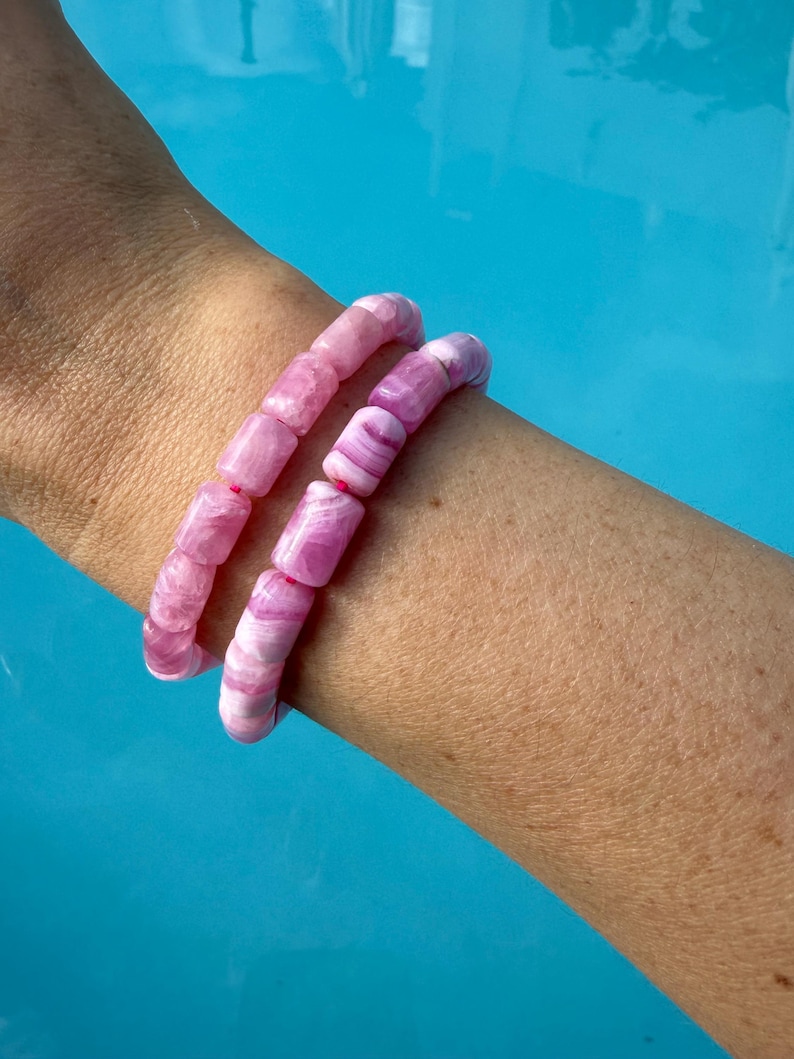 May include: Two pink beaded bracelets with a marbled effect are worn on a person's arm. The bracelets are made of cylindrical beads in shades of pink and white. The background is a bright blue.