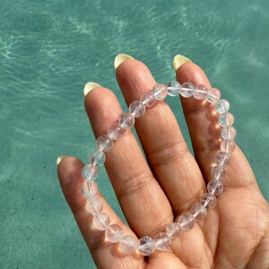 May include: A clear quartz crystal beaded bracelet. The bracelet is made of round, translucent beads strung together. The bracelet is held in a hand against a turquoise blue water background.
