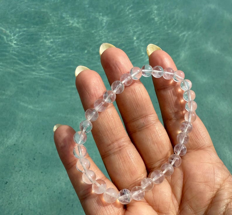 May include: A clear quartz crystal beaded bracelet. The bracelet is made of round, translucent beads strung together. The bracelet is held in a hand against a turquoise blue water background.