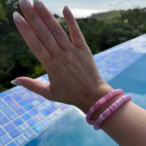 May include: Two pink beaded bracelets are worn on a wrist. The bracelets are made of rounded, irregular beads in shades of pink. The background includes a blue tiled surface and a natural outdoor setting with greenery and a cloudy sky.