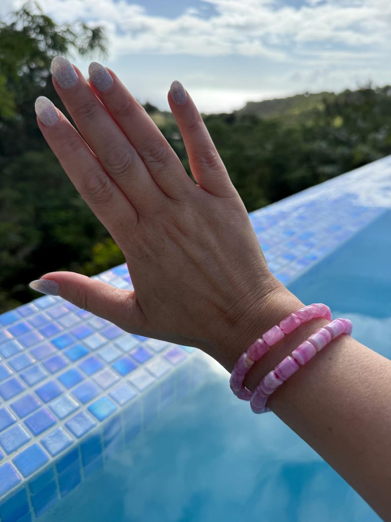 May include: Two pink beaded bracelets are worn on a wrist. The bracelets are made of rounded, irregular beads in shades of pink. The background includes a blue tiled surface and a natural outdoor setting with greenery and a cloudy sky.