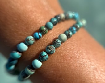 May include: Two turquoise beaded bracelets are shown. The bracelets are made of small, round beads in shades of turquoise, blue, and green. The bracelets are worn on a wrist.