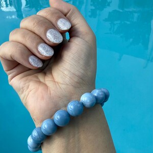 Puede incluir: Una pulsera de cuentas azul claro con cuentas grandes y redondas. La pulsera se lleva en una muñeca con esmalte de uñas plateado con purpurina. El fondo es azul brillante, posiblemente agua.
