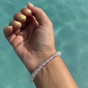 May include: A clear beaded bracelet is worn on a wrist with yellow painted nails. The bracelet is made of small, round, transparent beads. The background is a light blue, possibly water.