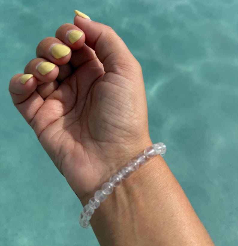 May include: A clear beaded bracelet is worn on a wrist with yellow painted nails. The bracelet is made of small, round, transparent beads. The background is a light blue, possibly water.