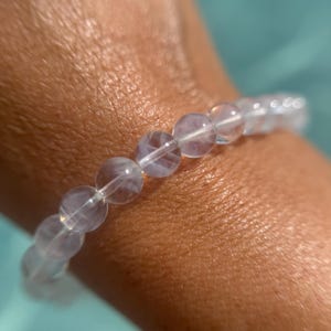 May include: A clear, beaded bracelet with a subtle, iridescent sheen. The round beads are strung together, creating a delicate and elegant accessory. The bracelet is shown against a soft, blurred background.