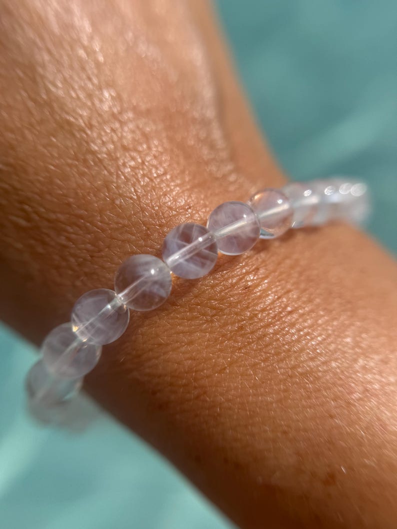 May include: A clear, beaded bracelet with a subtle, iridescent sheen. The round beads are strung together, creating a delicate and elegant accessory. The bracelet is shown against a soft, blurred background.