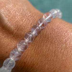 May include: A clear quartz beaded bracelet. The bracelet features translucent, round beads strung together on a white elastic cord. The beads have internal veining, creating a unique pattern. The bracelet is worn on a wrist.