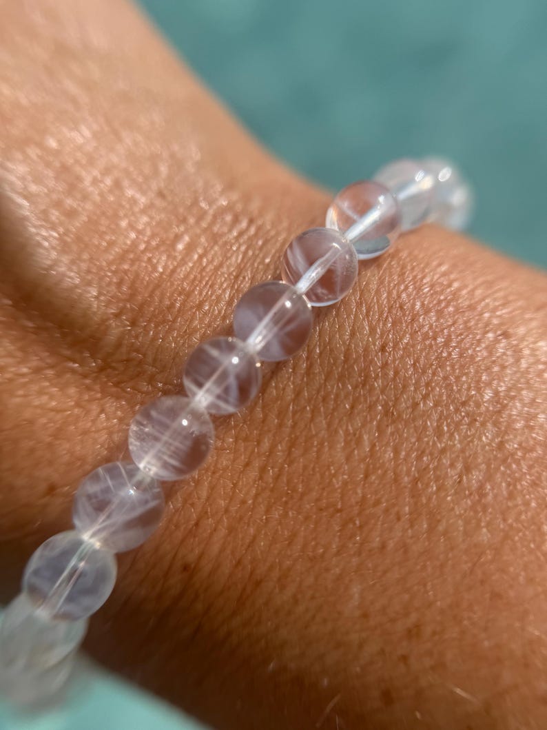 May include: A clear quartz beaded bracelet. The bracelet features translucent, round beads strung together on a white elastic cord. The beads have internal veining, creating a unique pattern. The bracelet is worn on a wrist.
