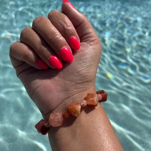 May include: A bracelet made of orange and brown carnelian gemstone beads. The beads are faceted and have a natural, rough texture. The bracelet is worn on a wrist with bright pink nail polish.