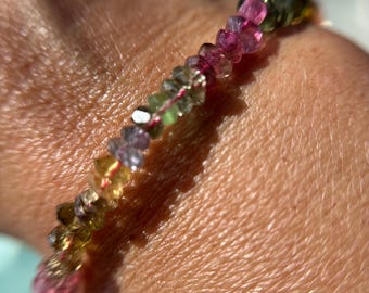 May include: A delicate bracelet composed of small, colorful gemstones. The stones are a mix of pink, green, yellow, and purple, strung together to form a flexible band. The bracelet is shown on a wrist.
