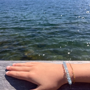 May include: A hand wearing a light blue beaded bracelet and a gold chain bracelet against a background of a lake and mountains.