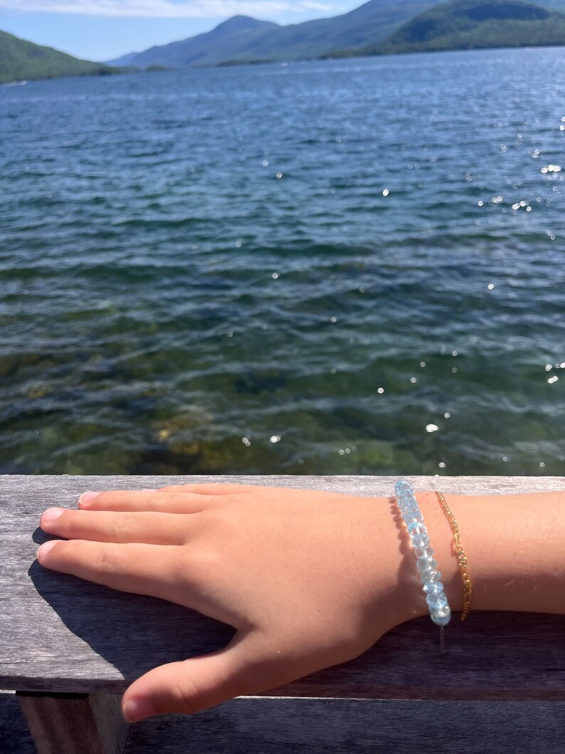 May include: A hand wearing a light blue beaded bracelet and a gold chain bracelet against a background of a lake and mountains.