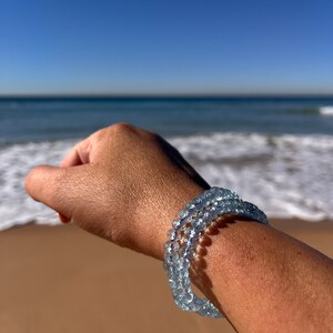 May include: A light blue aquamarine beaded bracelet with a delicate, layered design. The bracelet is worn on a person's wrist against a backdrop of a sandy beach and ocean waves.