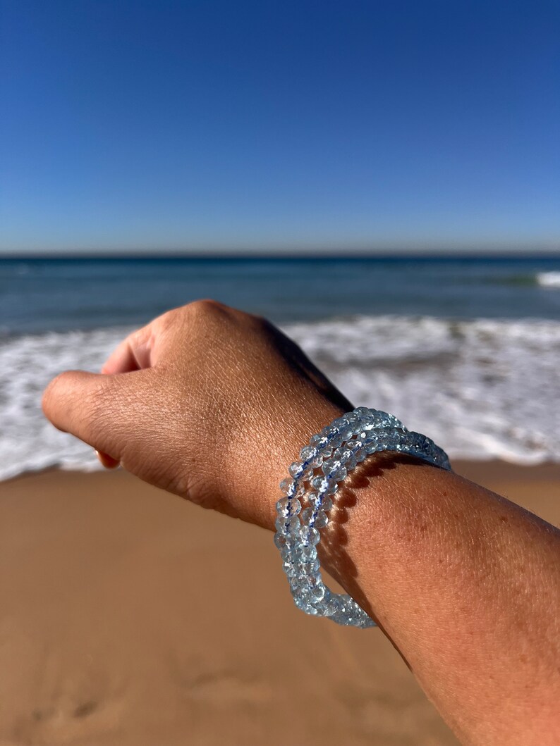 May include: A light blue aquamarine beaded bracelet with a delicate, layered design. The bracelet is worn on a person's wrist against a backdrop of a sandy beach and ocean waves.