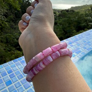 May include: Two pink and white beaded bracelets. The bracelets are made of rectangular beads with a marbled effect. The bracelets are stacked on top of each other. The background shows a pool with blue tiles and a view of trees and the ocean.