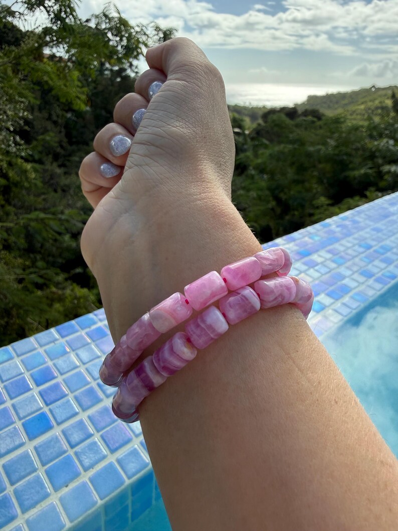 May include: Two pink and white beaded bracelets. The bracelets are made of rectangular beads with a marbled effect. The bracelets are stacked on top of each other. The background shows a pool with blue tiles and a view of trees and the ocean.