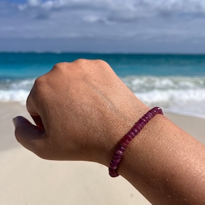 May include: A close-up of a person's wrist wearing a purple beaded bracelet. The bracelet is made of small, round beads that are strung together on a thin cord. The bracelet is worn on a tanned wrist against a background of white sand and blue water.