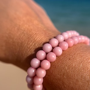 Könnte beinhalten: Ein Armband aus rosafarbenen Edelsteinen mit runden Perlen. Das Armband wird am Handgelenk getragen.