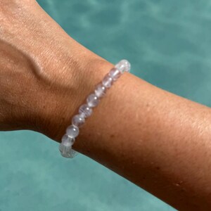 May include: A clear, beaded bracelet with a light purple hue is worn on a wrist. The bracelet is made of small, round beads strung together. The background is a blurred turquoise color.