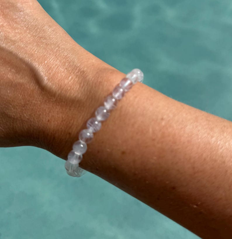 May include: A clear, beaded bracelet with a light purple hue is worn on a wrist. The bracelet is made of small, round beads strung together. The background is a blurred turquoise color.