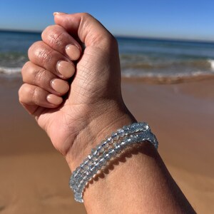 May include: A light blue crystal beaded bracelet on a person's wrist. The bracelet is made of small, round beads that are strung together on a thin cord. The beads are clear and have a slight sparkle to them.