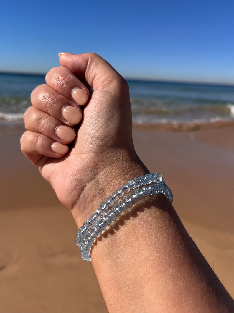 May include: A light blue crystal beaded bracelet on a person's wrist. The bracelet is made of small, round beads that are strung together on a thin cord. The beads are clear and have a slight sparkle to them.