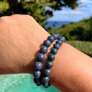 Könnte beinhalten: Zwei blaue Stein-Armbänder mit einem gesprenkelten Muster. Die Armbänder sind auf dem Handgelenk einer Person übereinander gelegt.