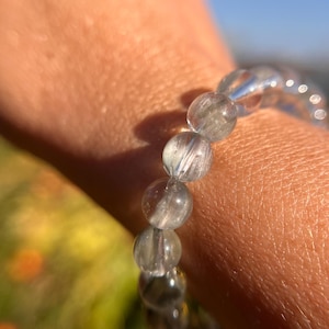 Puede incluir: Una pulsera de cuentas de cuarzo transparente con inclusiones plateadas. Las cuentas redondas están ensartadas, creando un accesorio delicado y elegante. La pulsera se muestra en una muñeca, con un fondo borroso verde y azul.