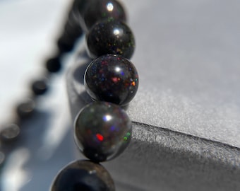 May include: A black opal beaded bracelet with iridescent rainbow colors. The beads are round and smooth, and the bracelet is lying on a gray surface.