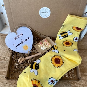 May include: A gift box with a yellow sock featuring bee and sunflower patterns, a wooden honey dipper, a heart-shaped ornament with the text "YOU ARE MY Sunshine", and a small decorative package.