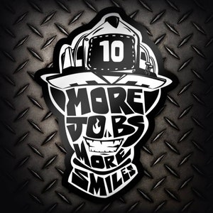 May include: Black and white sticker with a firefighter helmet design. The helmet has the number 10 on it. The text "More Jobs More Smiles" is written inside the helmet.