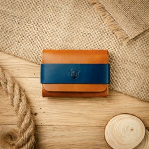 Peut inclure: Un portefeuille rectangulaire en cuir beige avec une sangle horizontale en cuir bleu foncé au centre. La sangle bleue est ornée d'un petit logo centré représentant la silhouette d'une tête de cerf à l'intérieur d'un contour en forme de bouclier.