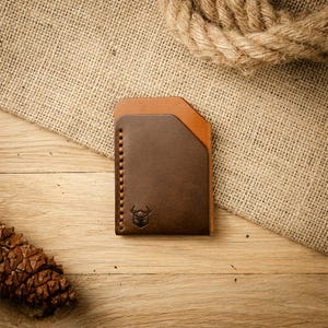 Peut inclure: Un porte-cartes en cuir bicolore composé d'un panneau avant marron foncé et d'un panneau arrière beige clair. Le portefeuille présente un bord supérieur incliné, des coutures contrastées visibles sur le côté gauche et un logo cerf embossé.