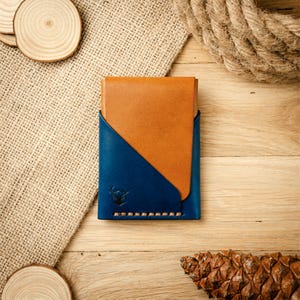 Peut inclure: Un portefeuille bi-fold bicolore composé d'une partie supérieure en cuir havane et d'une partie inférieure en cuir bleu foncé. La section bleue présente un petit logo de taureau embossé et des coutures apparentes de couleur claire à la base.