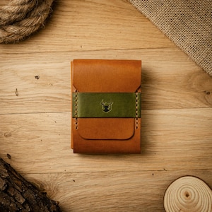 Peut inclure: Un portefeuille en cuir beige doté d'une bande horizontale en cuir vert olive au centre. La bande verte est ornée d'un petit logo en forme de tête de cerf foncé. Le portefeuille présente des coutures visibles sur les côtés de la bande.