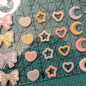 May include: Assortment of colorful iron-on patches in various shapes, including hearts, stars, moons, and bows. The patches are arranged on a blue cutting mat with white grid lines.