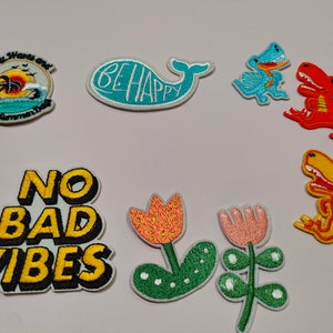 May include: Assortment of colorful embroidered iron-on patches featuring various designs, including a whale with the text "Be Happy", a dinosaur, a flower, and the phrase "No Bad Vibes".