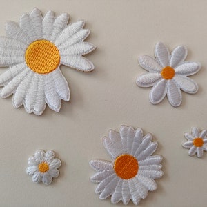 May include: Five white daisy flower iron-on patches with yellow centres. The patches are different sizes.