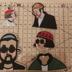 May include: Four embroidered patches featuring characters from the film Pulp Fiction. The patches are on a brown gridded surface. The characters are a man with a black hat and beard, a woman with a red hat and sunglasses, a man with a black hat and beard, and a woman with a white hat and a flower.