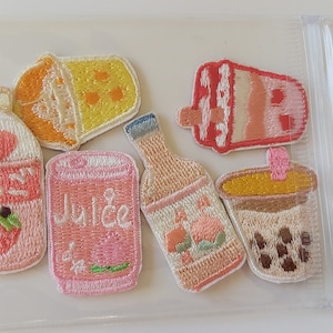 May include: A set of seven embroidered iron-on patches featuring various pink and yellow drinks, including a juice carton, a can of juice, a bottle of juice, a boba tea, and a cup of coffee. The patches are ideal for adding a touch of whimsy to clothing, bags, or other accessories.