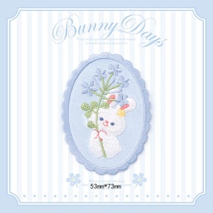 May include: A blue embroidered patch with a white bunny holding a bouquet of blue flowers. The patch is oval shaped with a scalloped edge. The text "Bunny Days" is written in a whimsical font above the patch. The text "If you hold a good intercourse with nature, it would teach you how to live." is written below the patch. The text "When we are out and about there 53mm*73mm of times" is written below the patch.