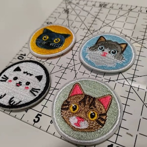 Peut inclure: Quatre patchs brodés thermocollants représentant différents chats de dessin animé. Les patchs sont circulaires et ont un fond blanc avec des contours noirs. Les chats ont des fourrures et des expressions faciales de différentes couleurs.