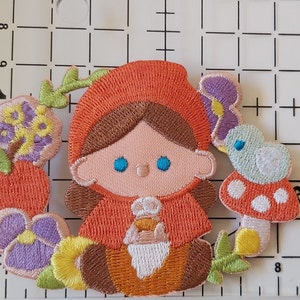 May include: Embroidered iron-on patch featuring a girl in a red hooded dress holding a basket of flowers. The patch is surrounded by flowers, a mushroom, and a bird.